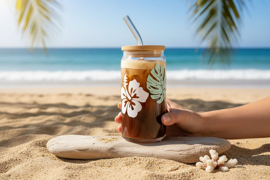 Between the Inks glass can tumbler with hibiscus and monstera leaf decals, bamboo lid, and glass straw on a beach.